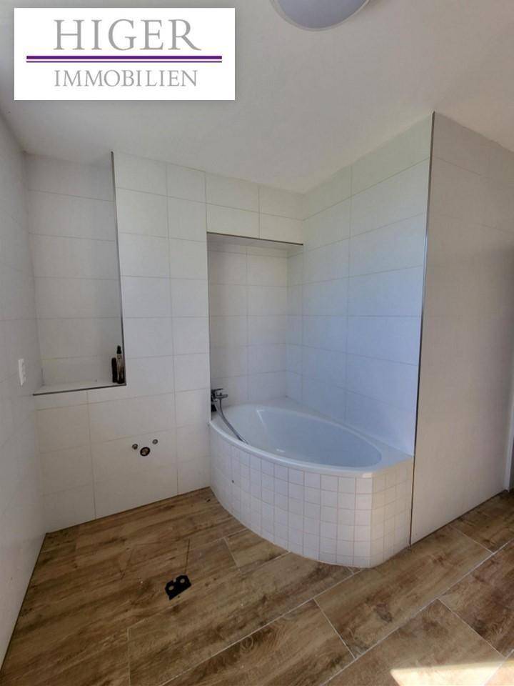 Badezimmer im Bau mit Eckbadewanne, weißen Fliesen an den Wänden und Holzoptik-Fliesenboden.