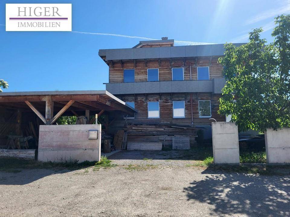 Das Holzgebäude mit einem angrenzenden Holzcarport und Baumaterialien auf dem Grundstück.