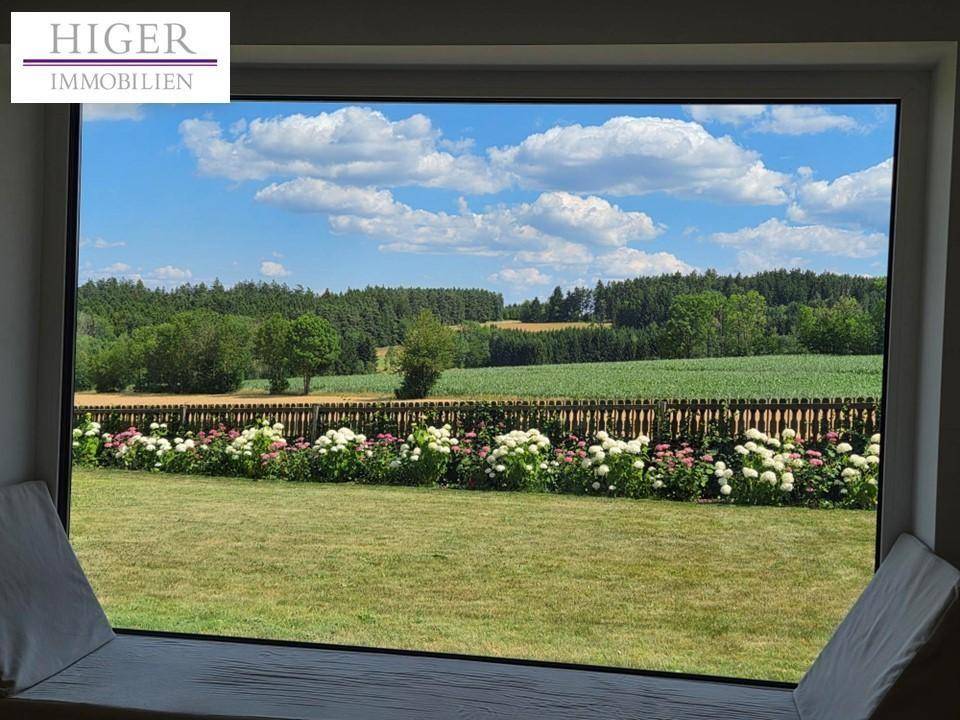 Große Fensterfront mit weitem Blick über grüne Felder und Wälder unter blauem Himmel.