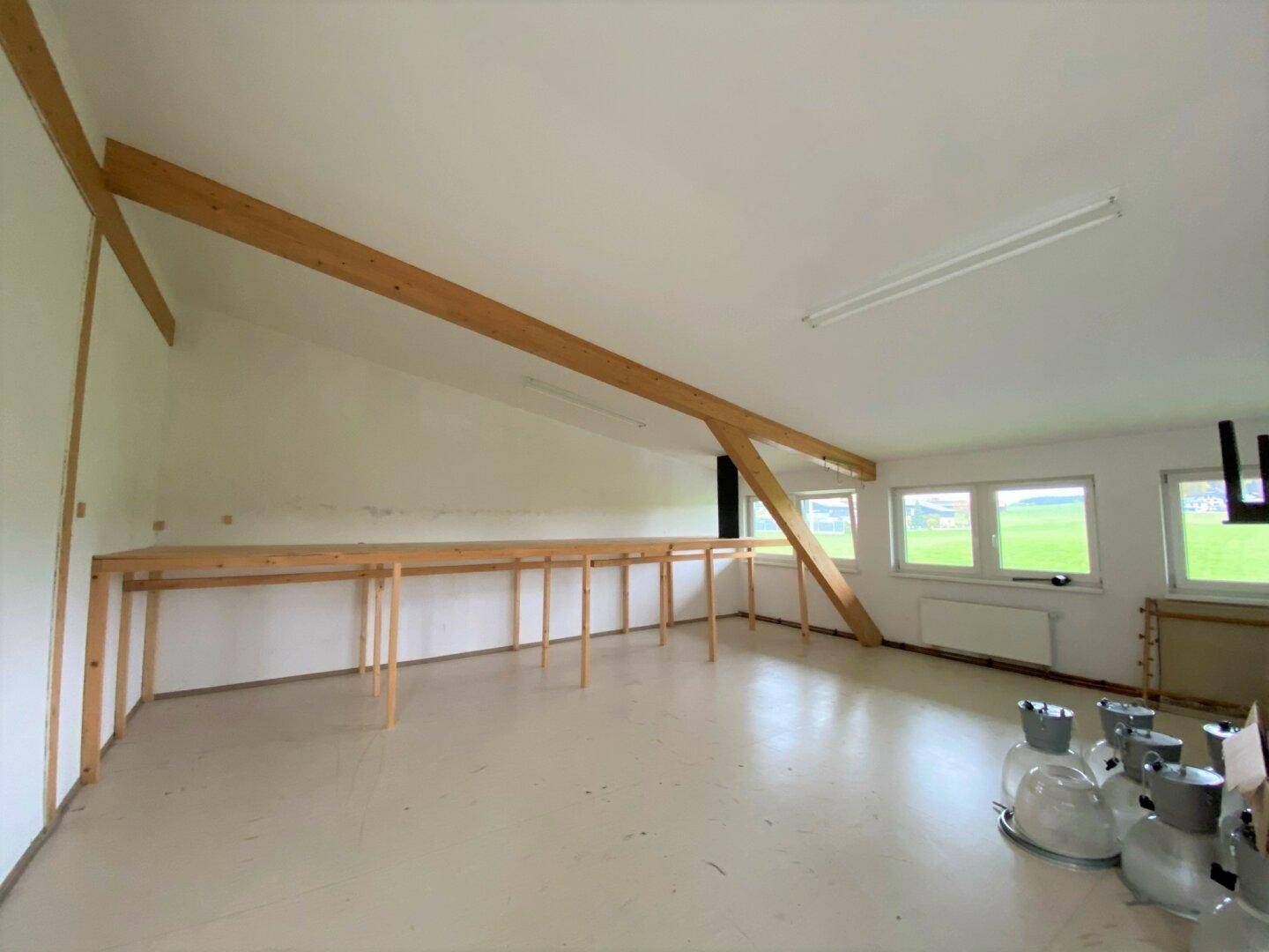 Heller Dachboden mit sichtbaren Holzbalken, großen Fenstern und langen Arbeitstischen mit Blick ins Grüne.