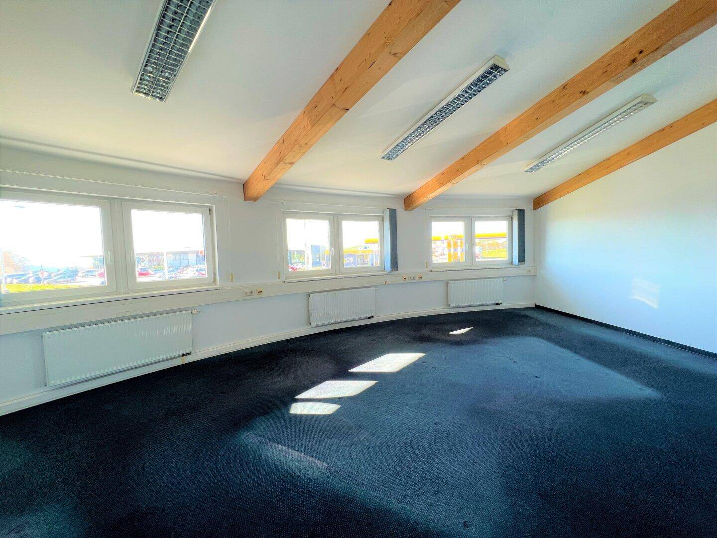 Heller Büroraum mit Teppichboden, sichtbaren Holzbalken und großen Fenstern, ideal für gewerbliche Nutzung.