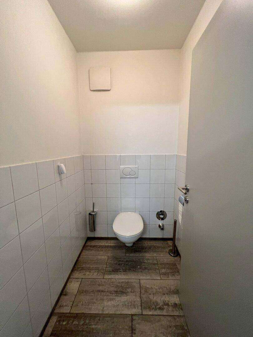 Kleines, helles Gäste-WC mit weißen Fliesen, Holzbodenoptik und Wand-WC, ausgestattet mit einem Abluftventilator.