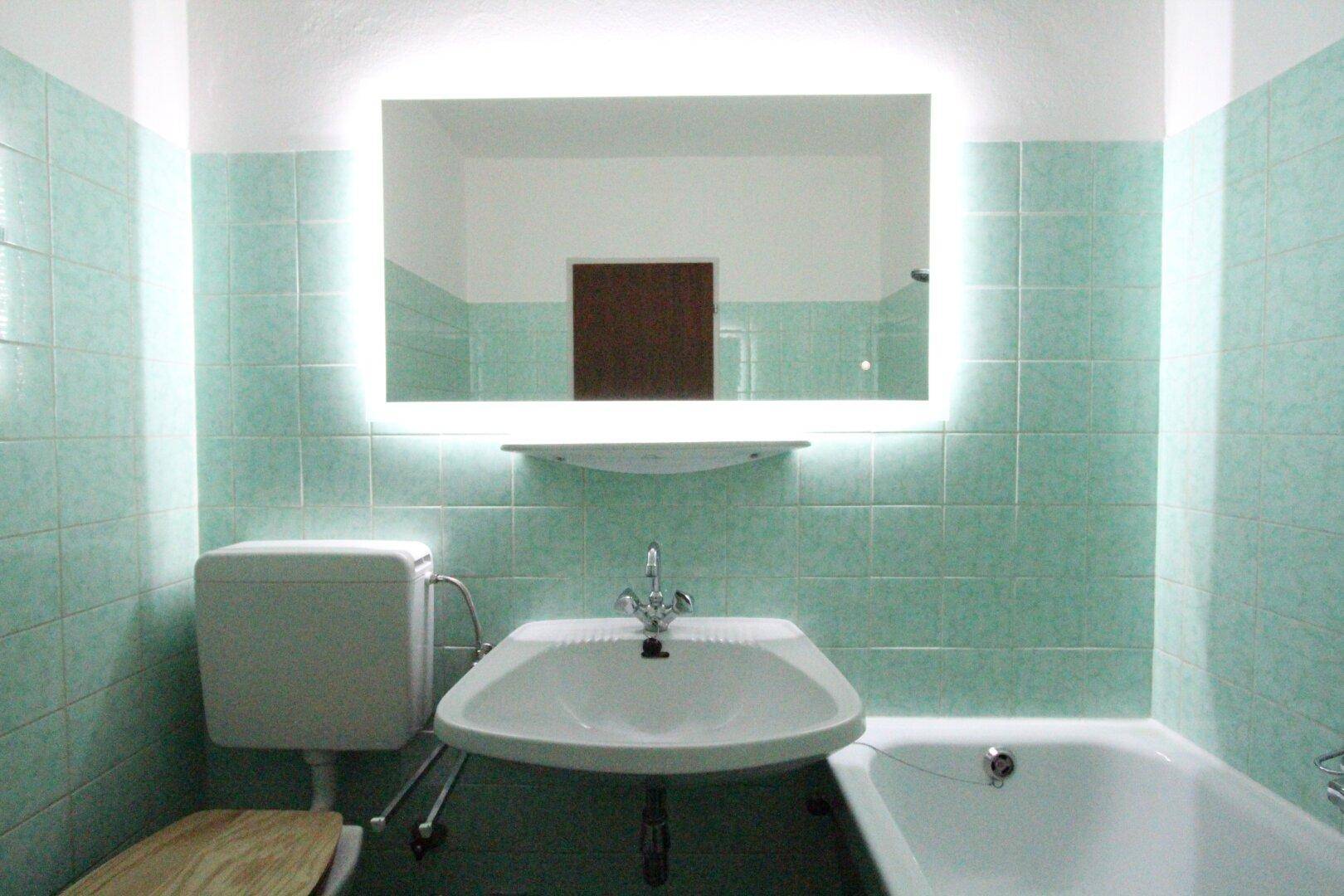 Badezimmer mit mintgrünen Fliesen, Waschbecken, Toilette und Badewanne, beleuchteter Spiegel.
