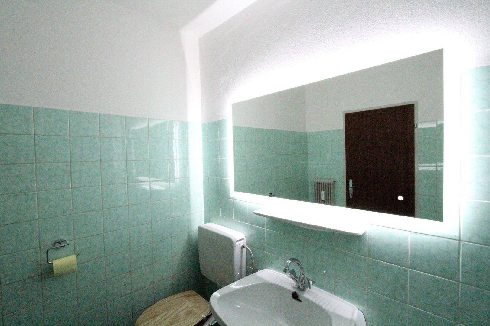 Badezimmer mit WC, Waschbecken und einem großen, beleuchteten Spiegel über den mintgrünen Fliesen.