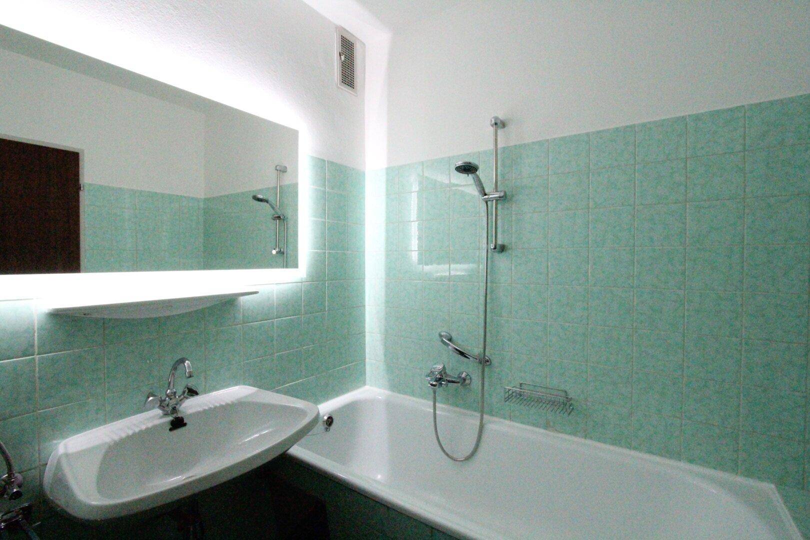 Badezimmer mit Badewanne inklusive Duschfunktion, Waschbecken und einem modernen beleuchteten Spiegel.