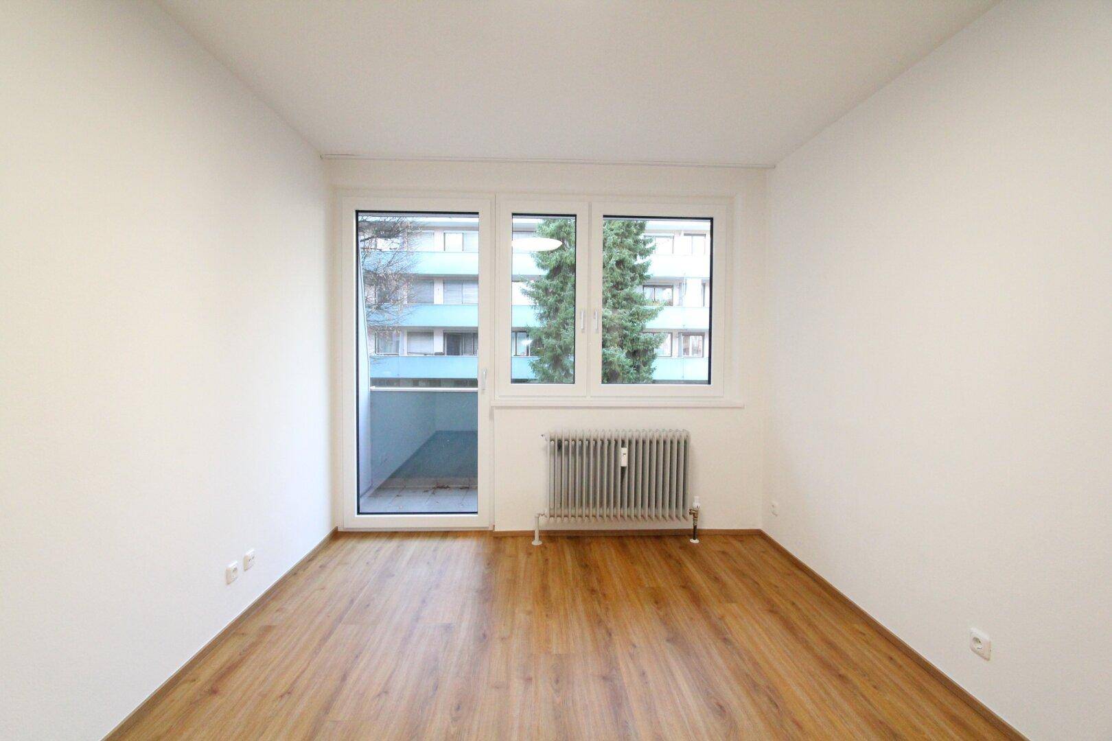 Geräumiges Zimmer mit Laminatboden und großem Fenster, das viel Tageslicht hereinlässt.