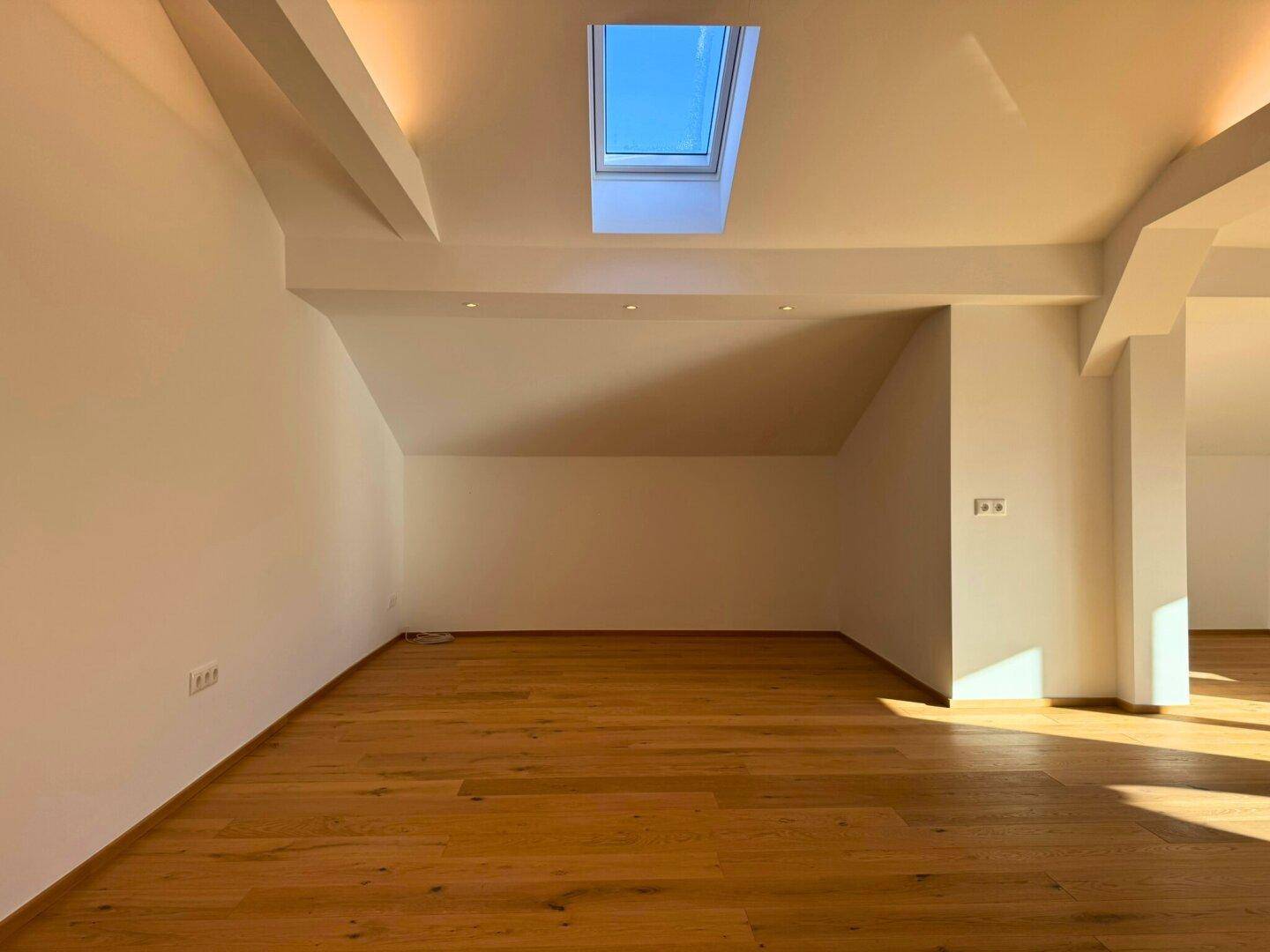 Leerer Wohnbereich mit Holzboden, Dachschrägen und einem hellen Dachfenster.