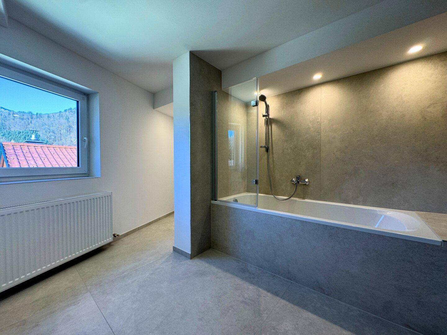 Modernes Badezimmer mit Badewanne, Duschwand, großen Fliesen und Fenster.