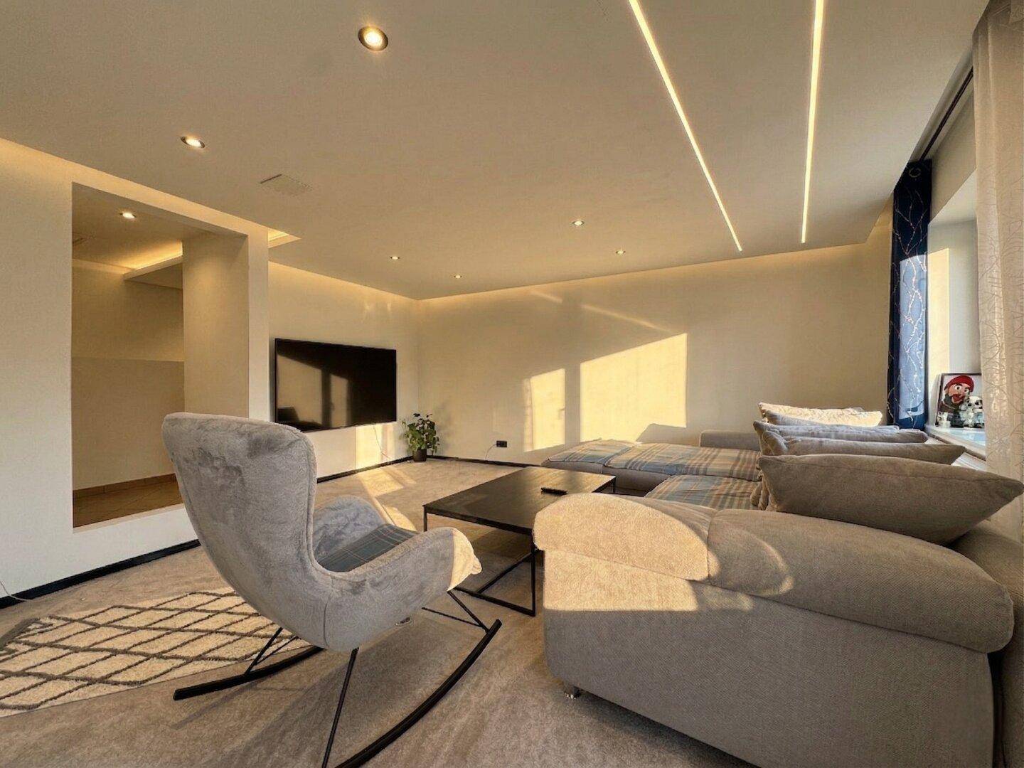 Modernes Wohnzimmer mit großem Sofa, indirekter Beleuchtung und viel natürlichem Licht durch die Fenster.