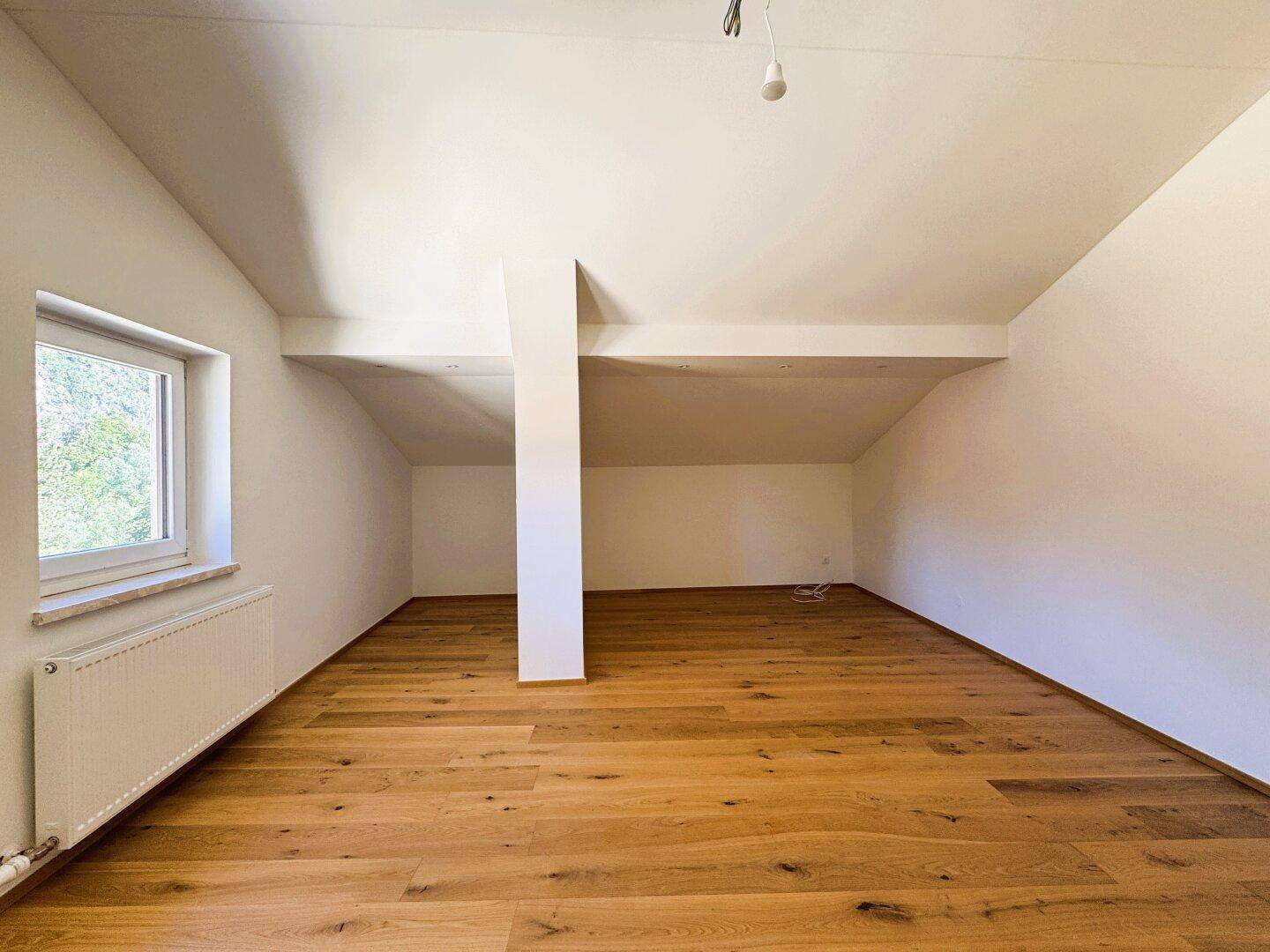 Helles Zimmer mit Holzboden, Dachschräge und Fenster mit Blick ins Grüne.