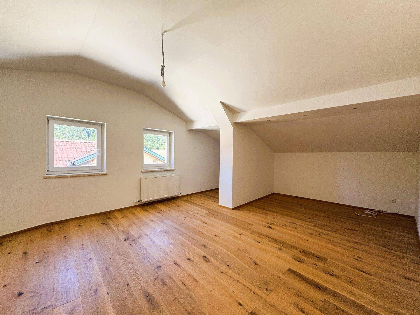 Geräumiges Zimmer mit Holzboden, Dachschräge und zwei Fenstern für viel Tageslicht.