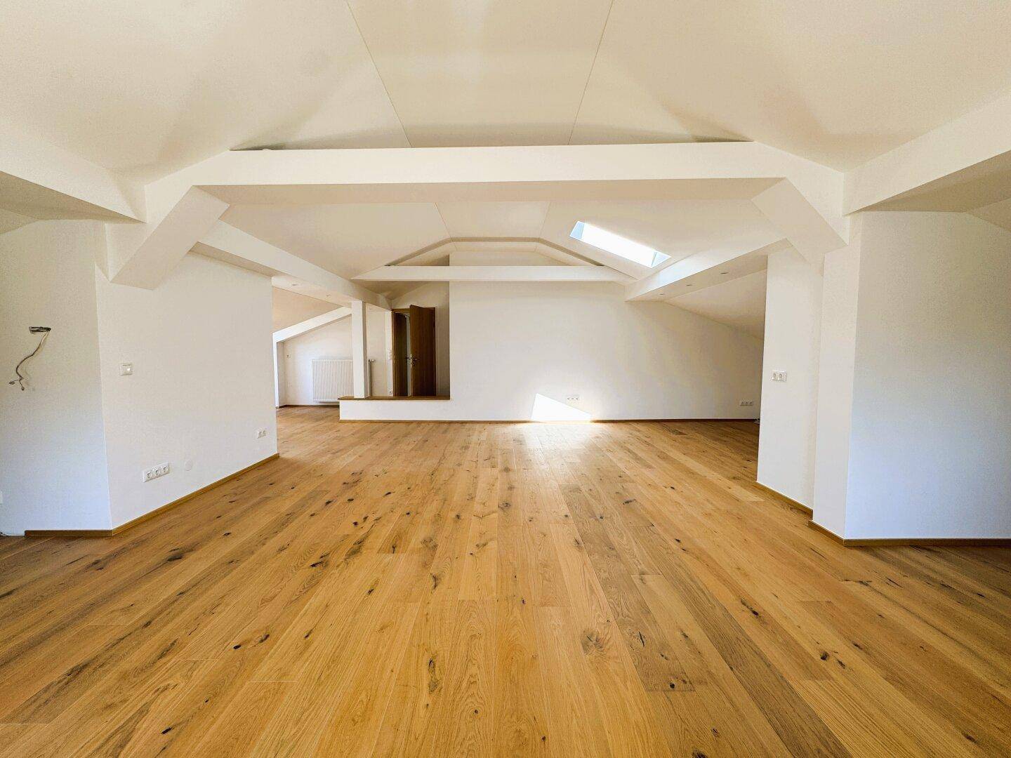 Weitläufiger Dachgeschossraum mit sichtbaren Balken, Holzboden und viel natürlichem Licht.