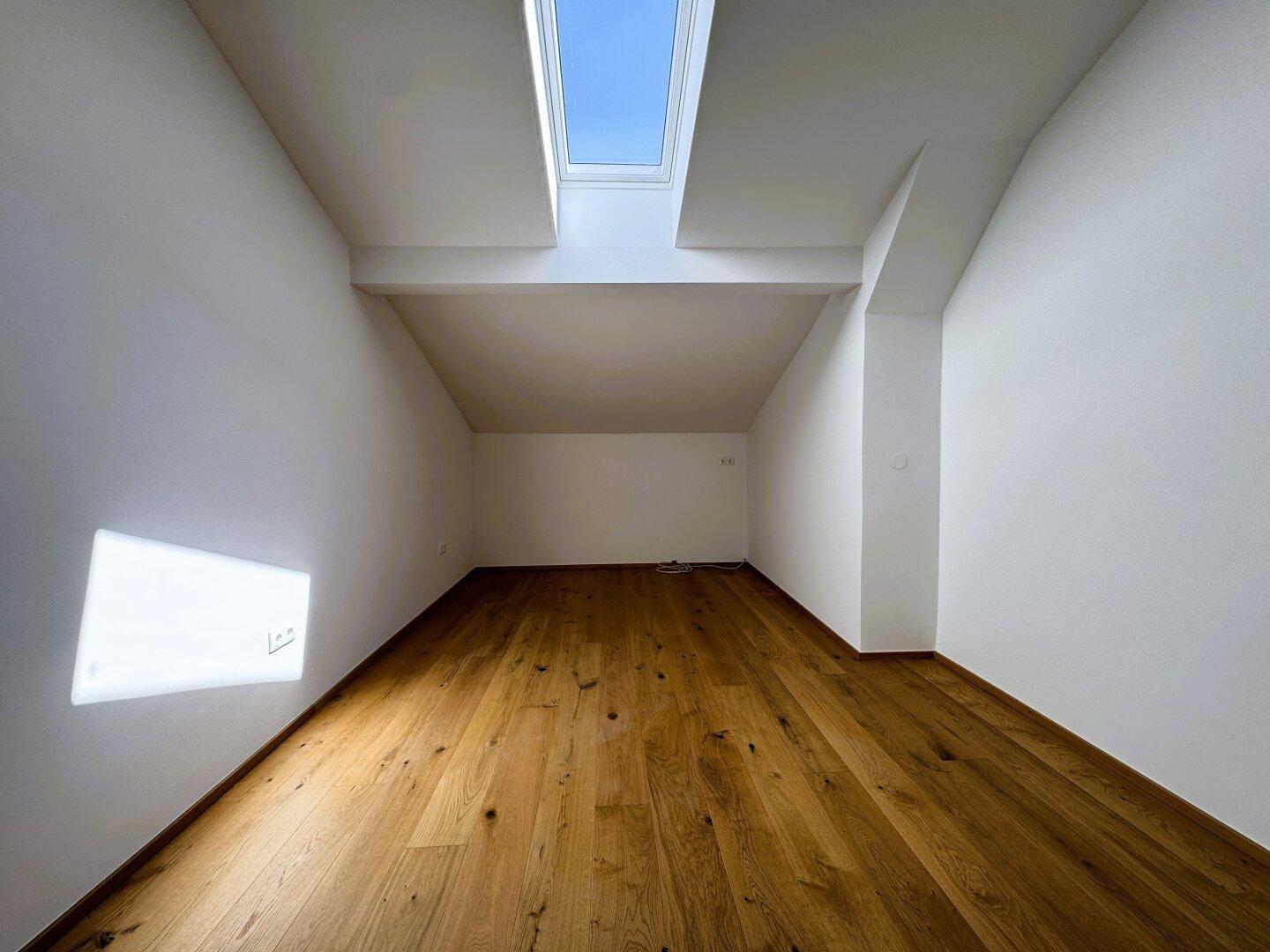 Schmaler Raum mit Holzboden, Dachschräge und einem Dachfenster für natürliches Licht.