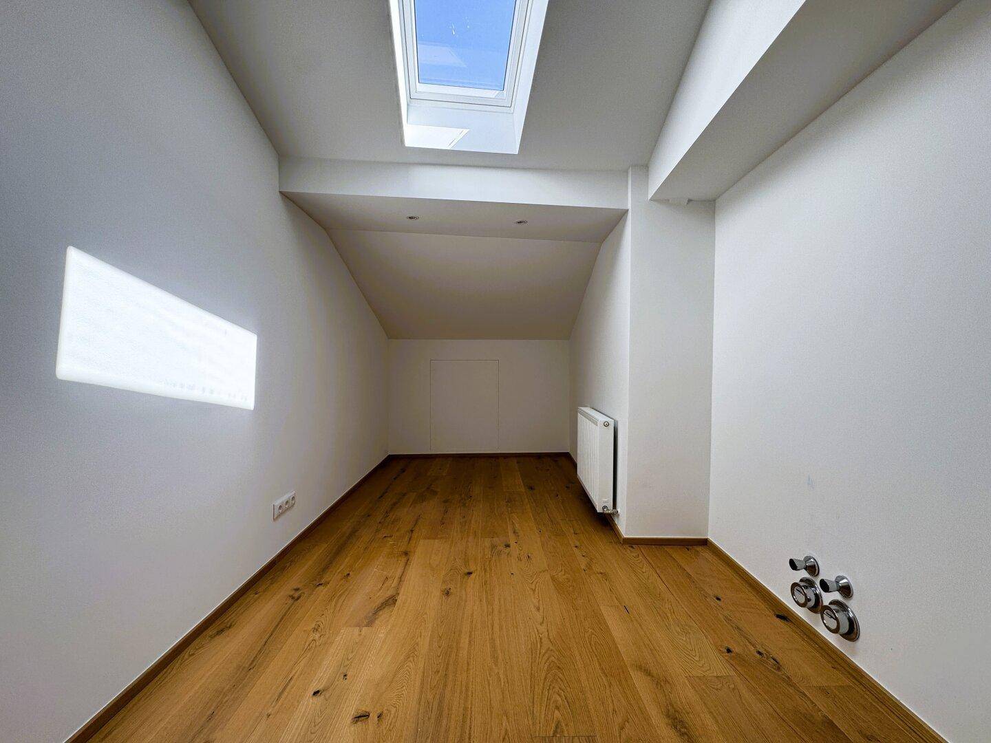 Heller Raum mit Holzboden, Dachschräge, Dachfenster und Heizkörper.