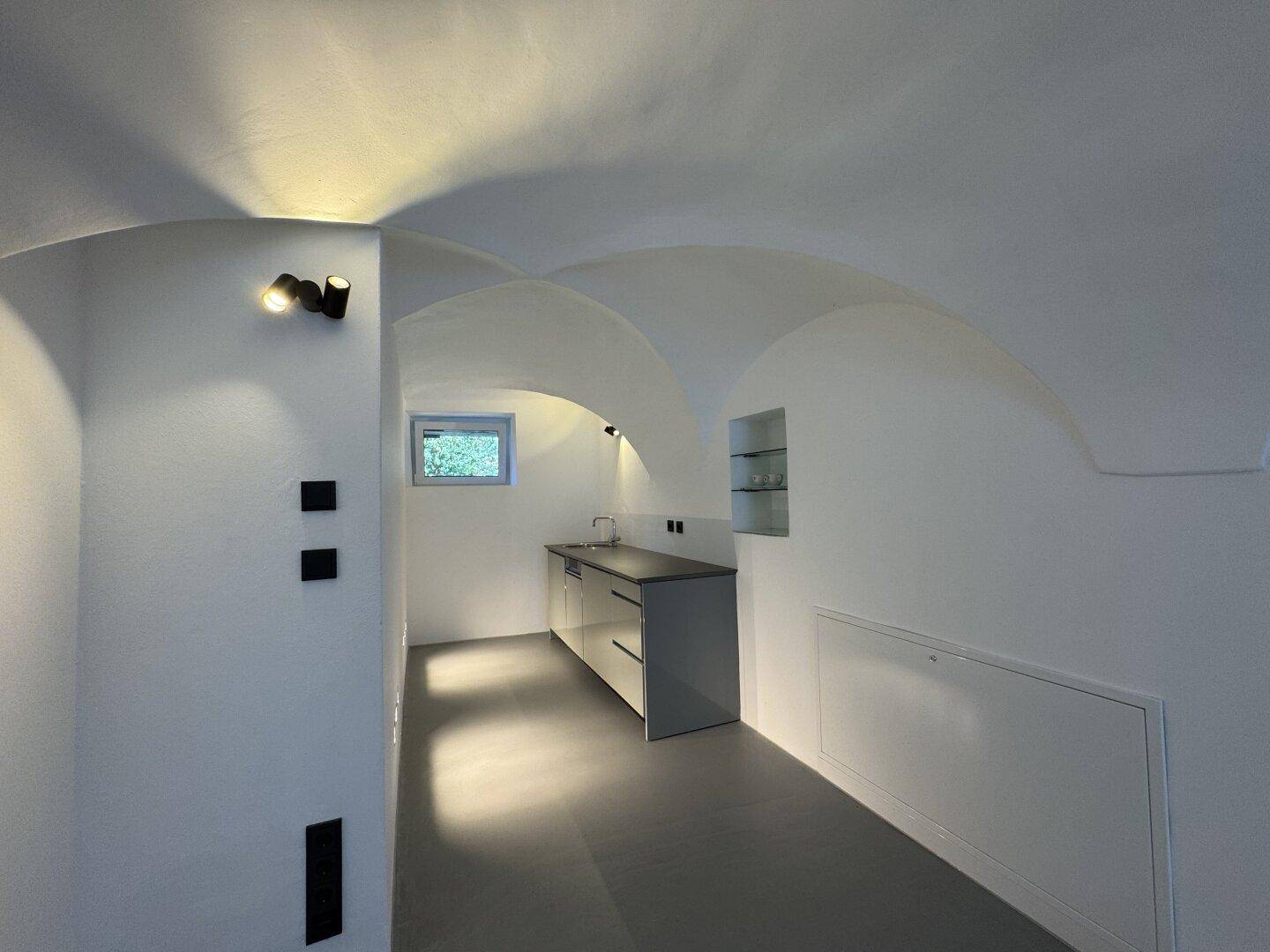 Kleine Küchenzeile mit Spüle, Unterschränken und einem Fenster in einem Raum mit Gewölbedecke.