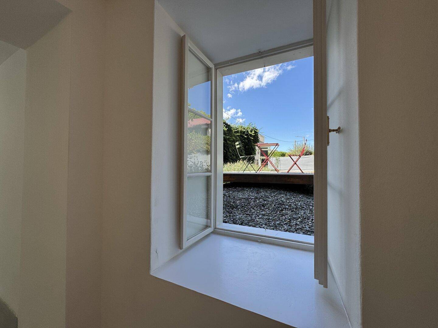 Offenes Fenster mit Blick auf eine Außenterrasse mit Holzboden und Sitzgelegenheiten unter blauem Himmel.