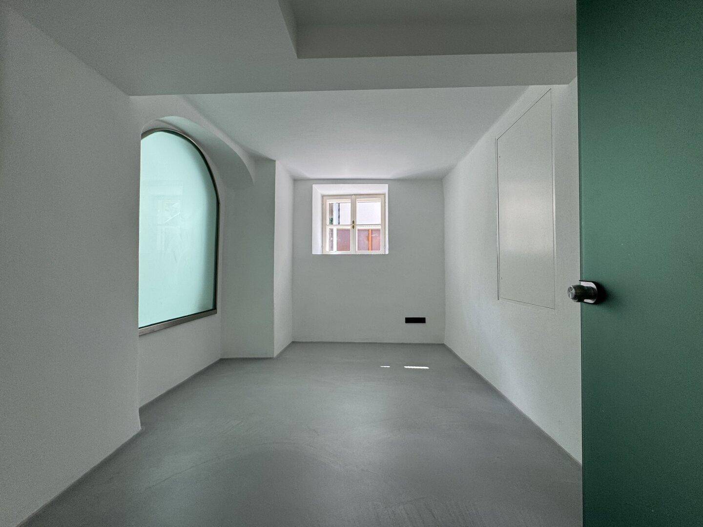 Leerer Raum mit hellen Wänden, modernem Boden und zwei Fenstern, darunter ein markantes Rundbogenfenster.