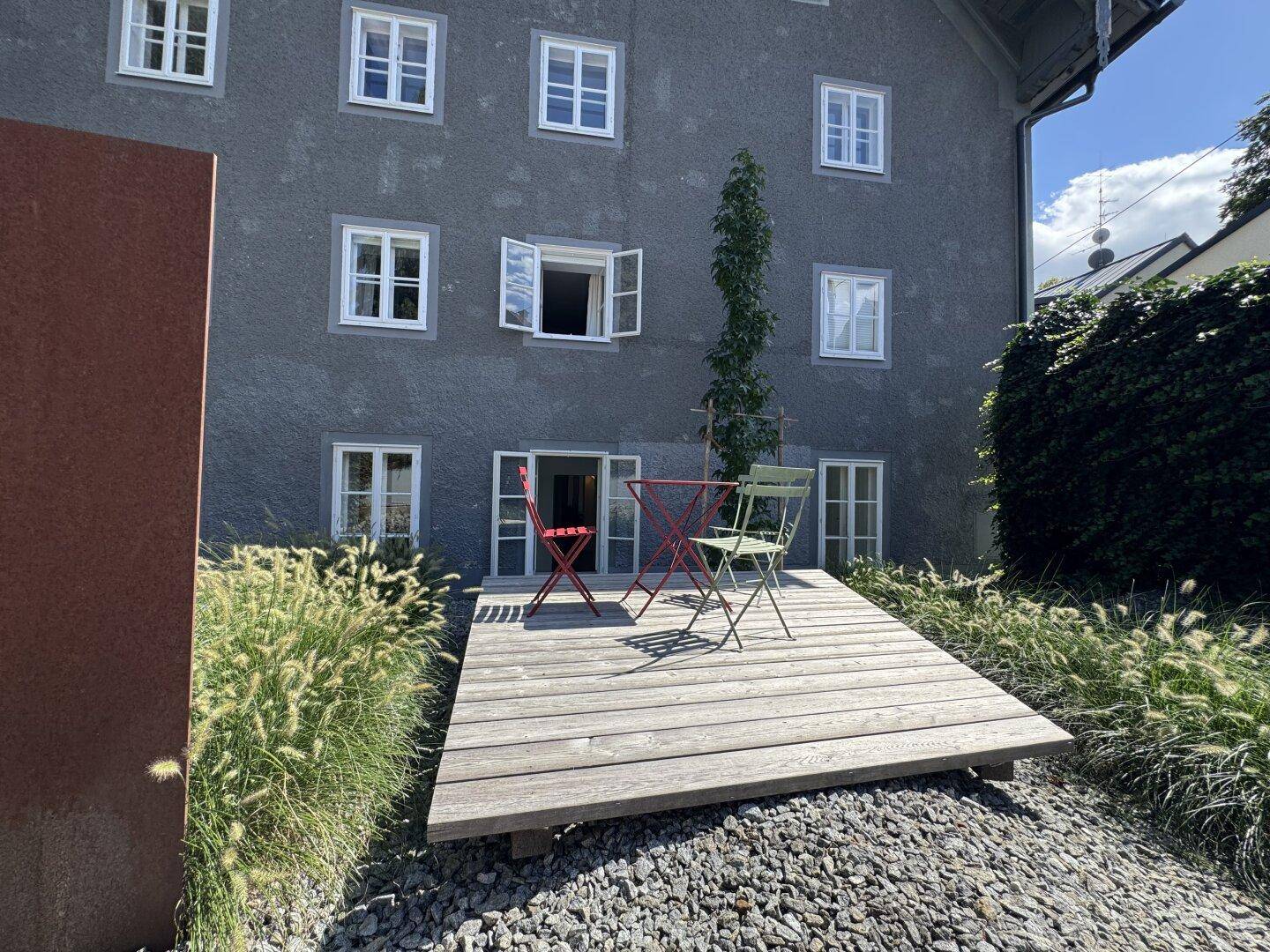 Außenbereich mit Holzterrasse, Gartenmöbeln und Blick auf die graue Fassade des Gebäudes.