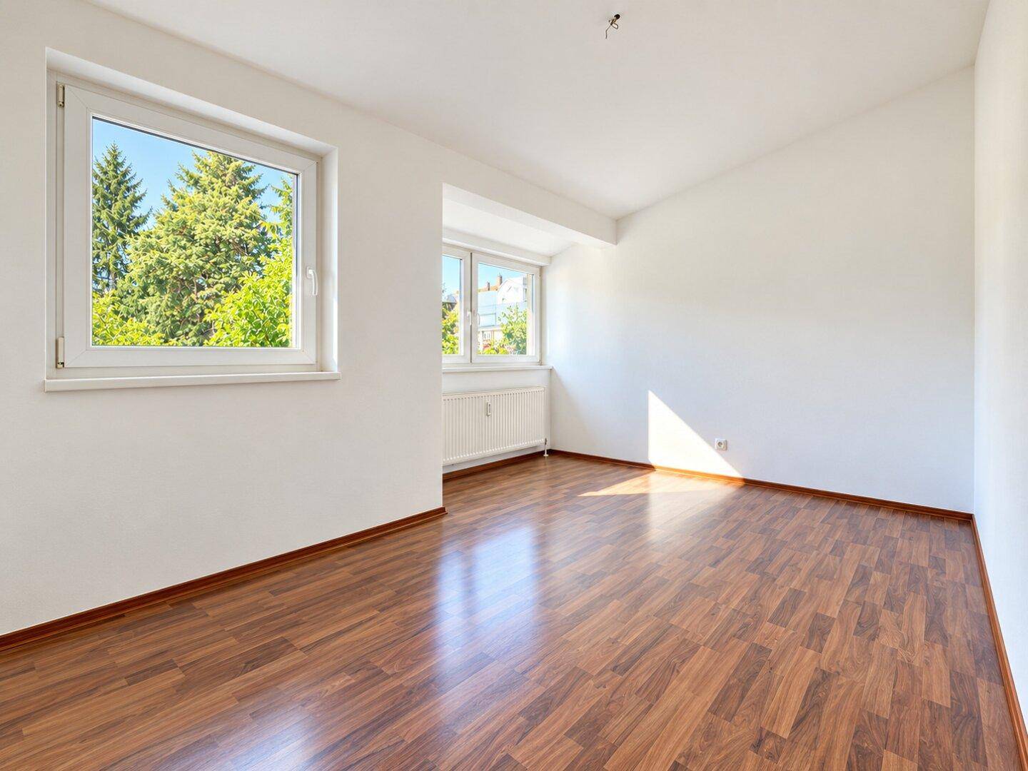 Helles Zimmer mit Laminatboden und zwei Fenstern, die viel Tageslicht hereinlassen.