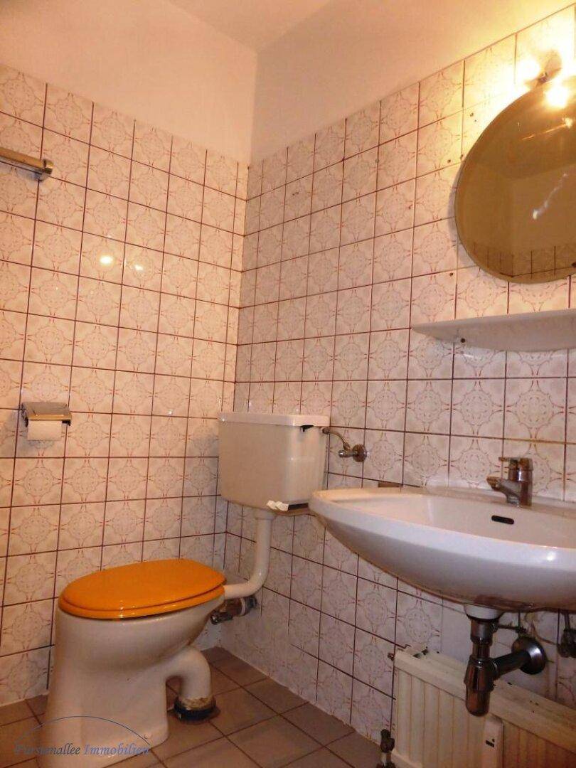 Badezimmer mit karierten Fliesen, WC mit orangefarbenem Sitz, Waschbecken und einem runden Spiegel.