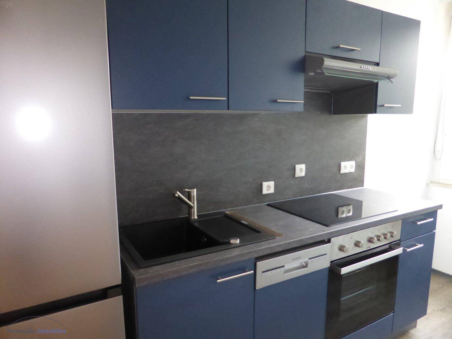 Moderne Küche mit blauen Schränken, dunkler Arbeitsplatte und integrierten Geräten, inklusive Geschirrspüler und Induktionskochfeld.