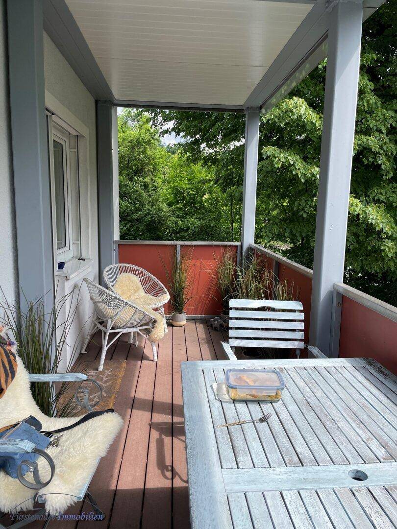 Gemütlicher Balkon mit Holzboden, bequemen Sitzgelegenheiten und viel Grün im Hintergrund.