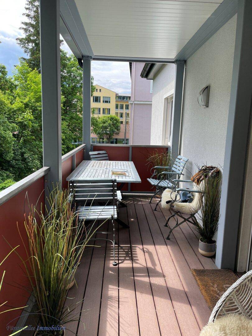Überdachter Balkon mit Holzboden, Esstisch und Stühlen, umgeben von üppigem Grün und Bäumen.