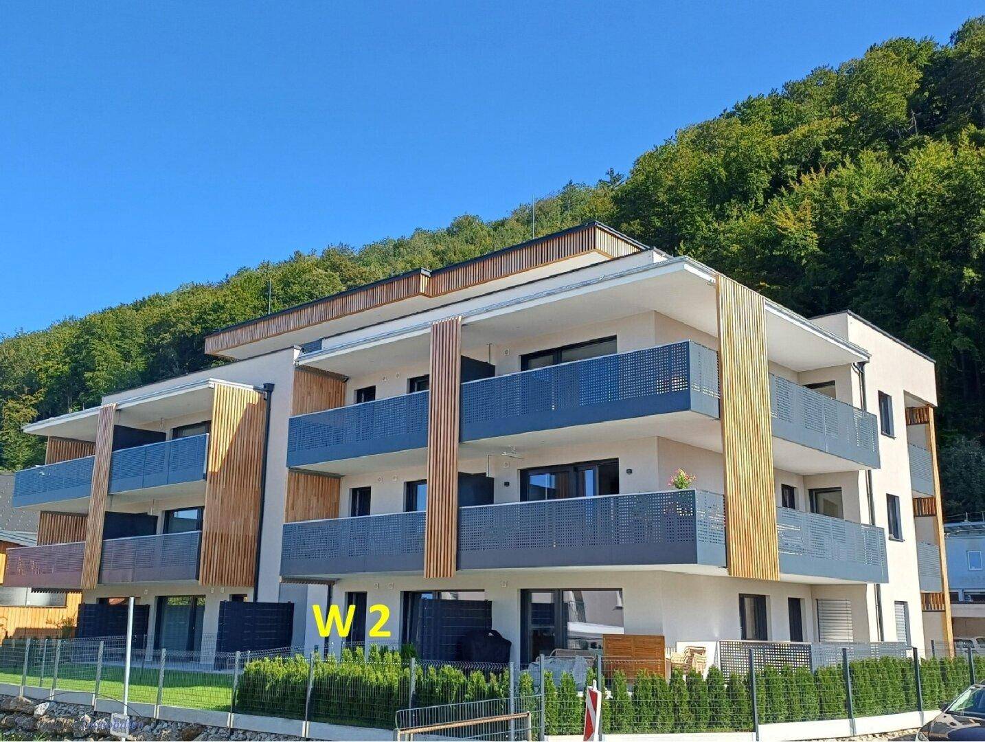 Modernes Mehrfamilienhaus mit Holzverkleidung, großen Balkonen und Flachdach vor bewaldetem Hügel.