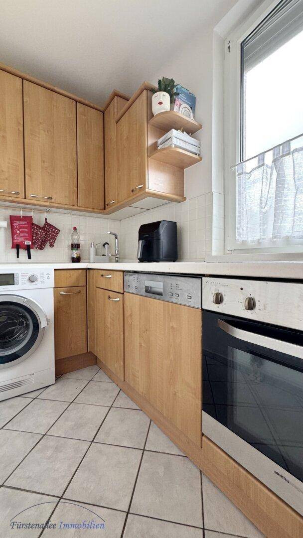 Funktionale Küche mit Holzschränken, Fliesenboden, Spülmaschine, Backofen und Fenster.