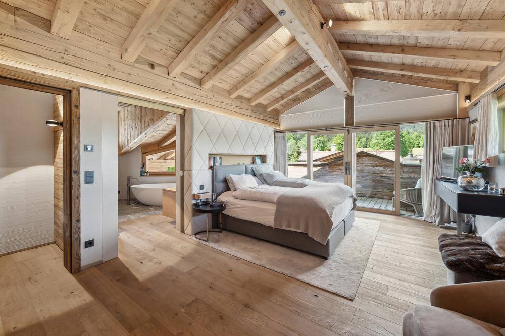 Gemütliches Schlafzimmer mit Holzbalken, Zugang zum Balkon und freistehender Badewanne im offenen Bereich.