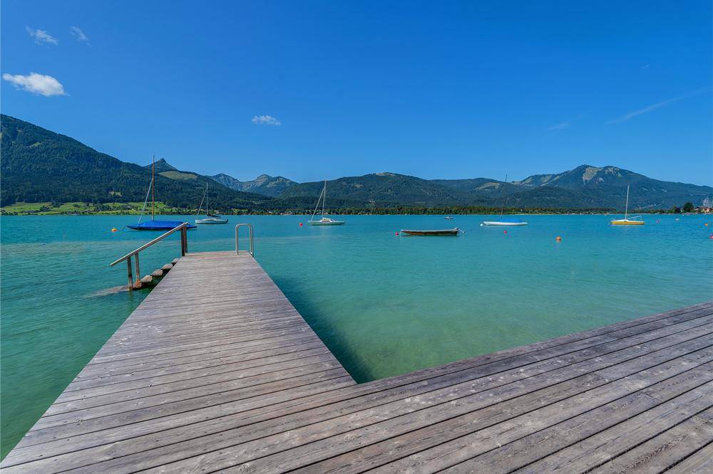 Holzsteg am klaren, türkisfarbenen See mit Segelbooten und umliegenden Bergen unter blauem Himmel.