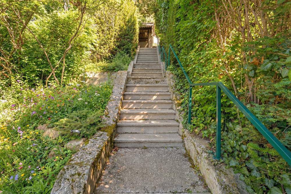 Steile Steintreppe mit Geländer, umgeben von üppigem Grün und Blumenbeeten.