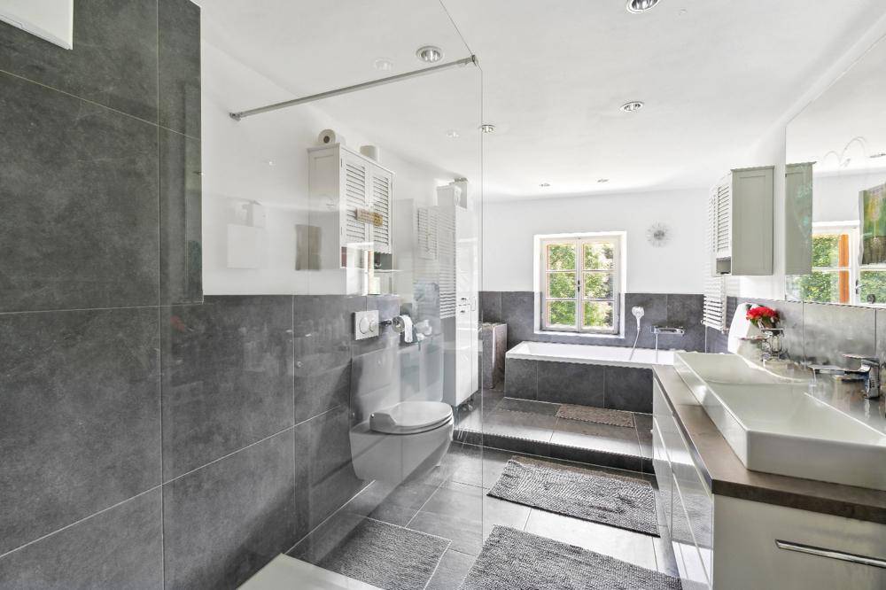 Modernes Badezimmer mit grauen Fliesen, Doppelwaschbecken, begehbarer Dusche und Badewanne am Fenster.