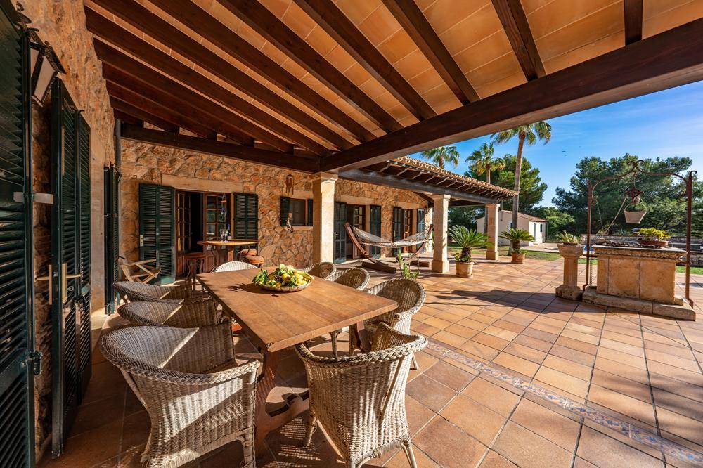 Große überdachte Terrasse mit Holzbalken, Esstisch und Korbstühlen, ideal für Mahlzeiten im Freien.