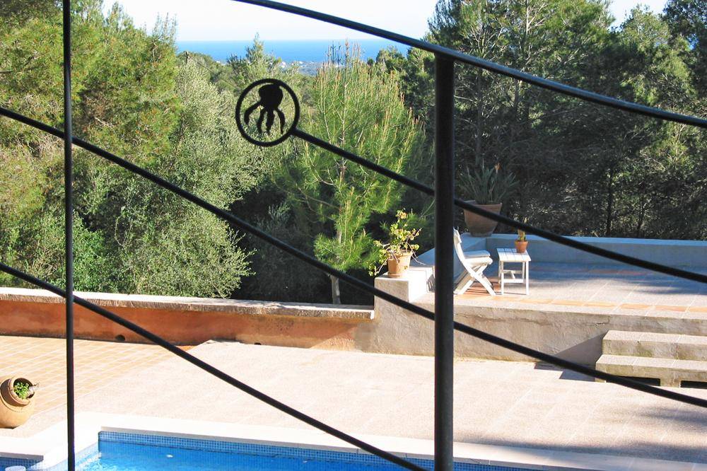 Blick von einer Terrasse mit schmiedeeisernem Geländer auf den Pool, Bäume und das ferne Meer.