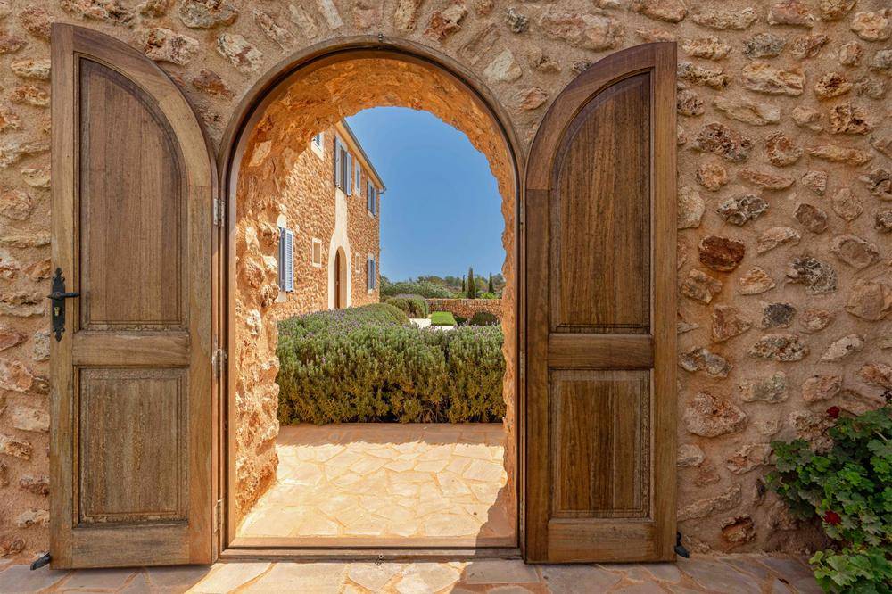 Rustikaler Steinbogen führt zu einem gepflegten Gartenbereich mit mediterraner Bepflanzung und Villa im Hintergrund.
