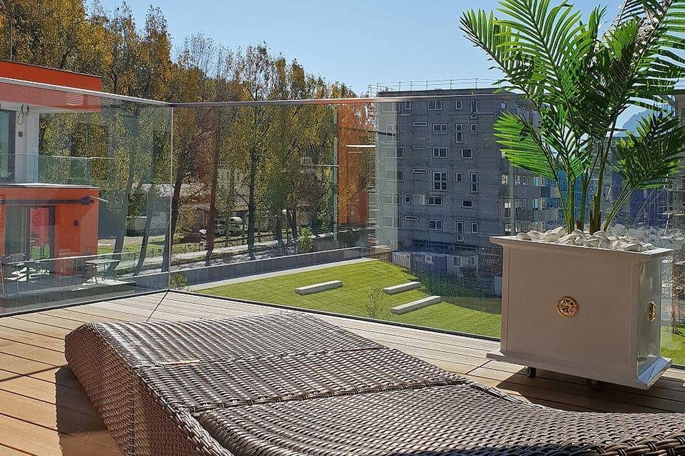Geräumiger Balkon mit Holzdeck, Glasgeländer und Liegestuhl, umgeben von Grünflächen.