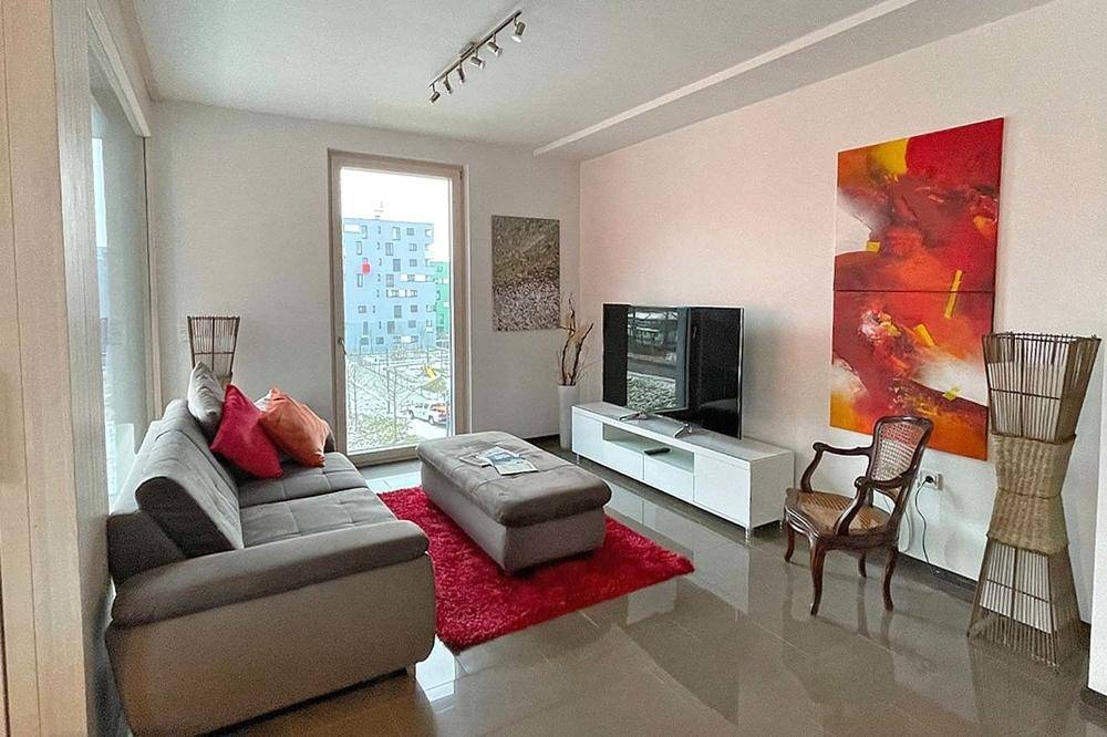 Helles Wohnzimmer mit beigem Sofa, rotem Teppich und Blick auf die umliegenden Gebäude.