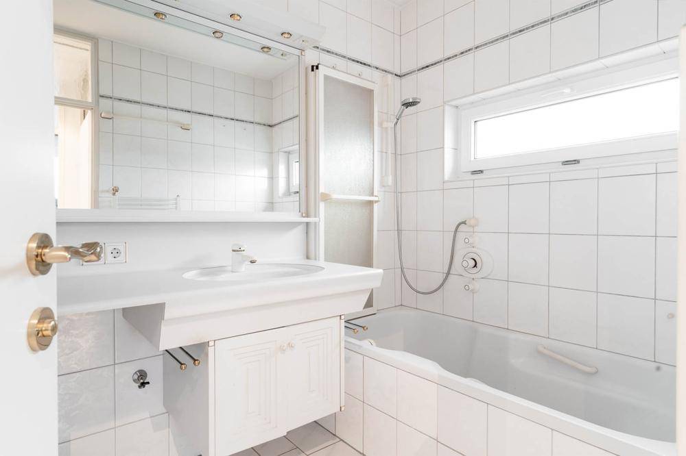 Helles Badezimmer mit weißen Fliesen, Badewanne mit Duschfunktion und einem Fenster.