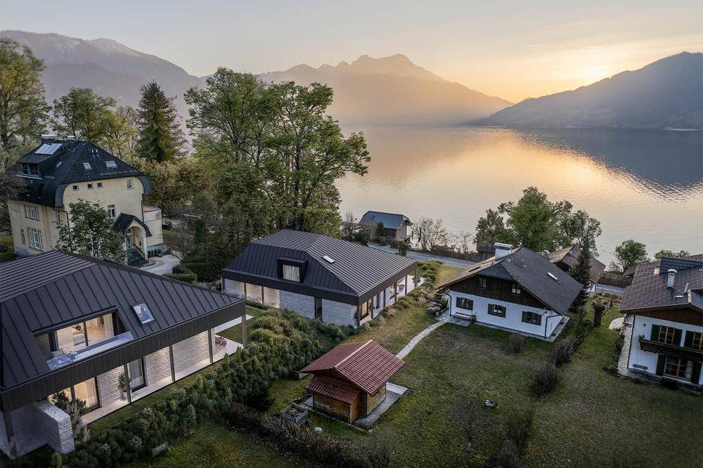 Panoramablick auf mehrere Häuser am Seeufer mit Bergen im Hintergrund bei Sonnenuntergang.