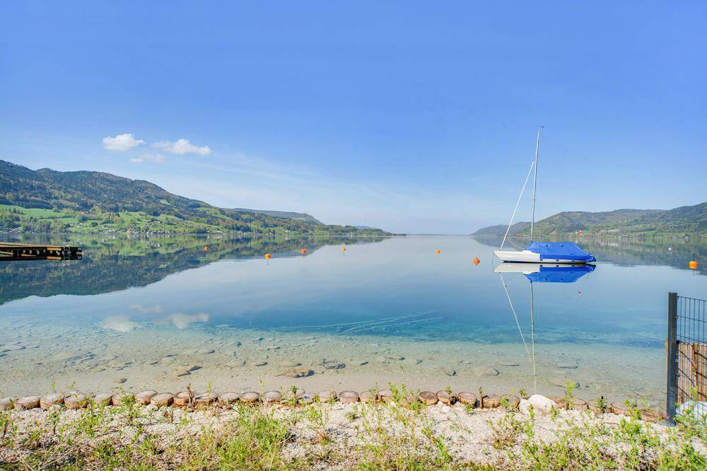Ruhiger See mit Segelboot und klarem Wasser, umgeben von sanften Hügeln und Bergen.