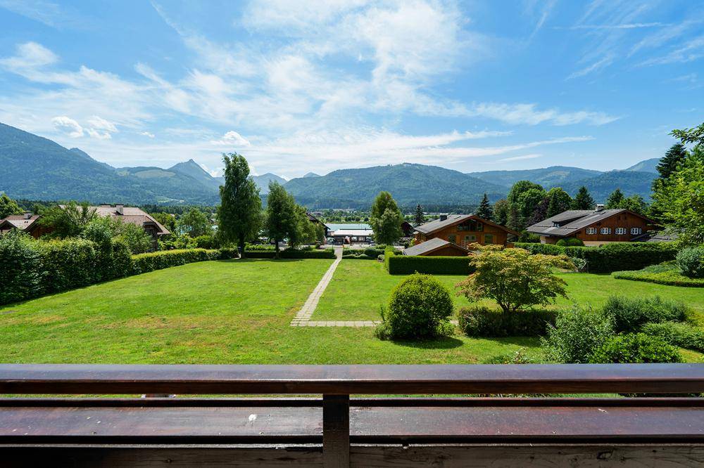 Weitläufiger Blick über eine grüne Wiese und Häuser auf die umliegenden Berge.