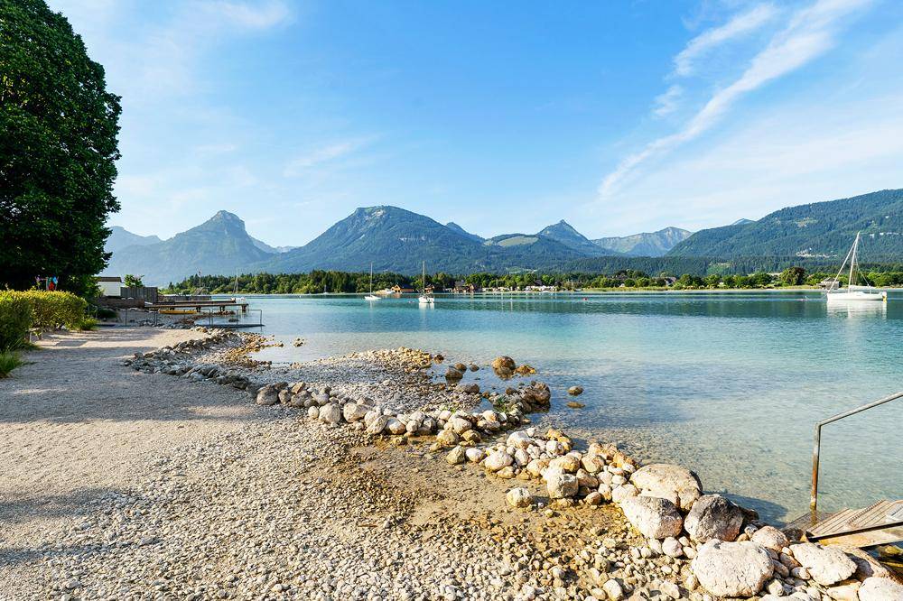 Klarer See mit Kiesstrand und Bergen im Hintergrund unter blauem Himmel.