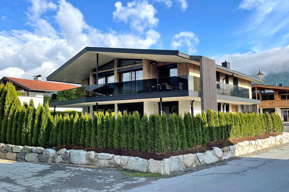 Elegante moderne Villa mit Holzfassade und gepflegter Hecke in alpiner Umgebung.