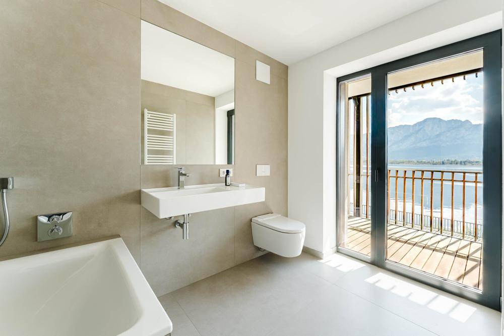 Modernes Badezimmer mit Badewanne, wandhängendem WC und Balkonzugang mit Seeblick.