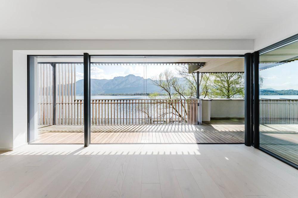 Heller Innenraum mit großen Glasfronten, die sich zu einem Balkon mit See- und Bergblick öffnen.