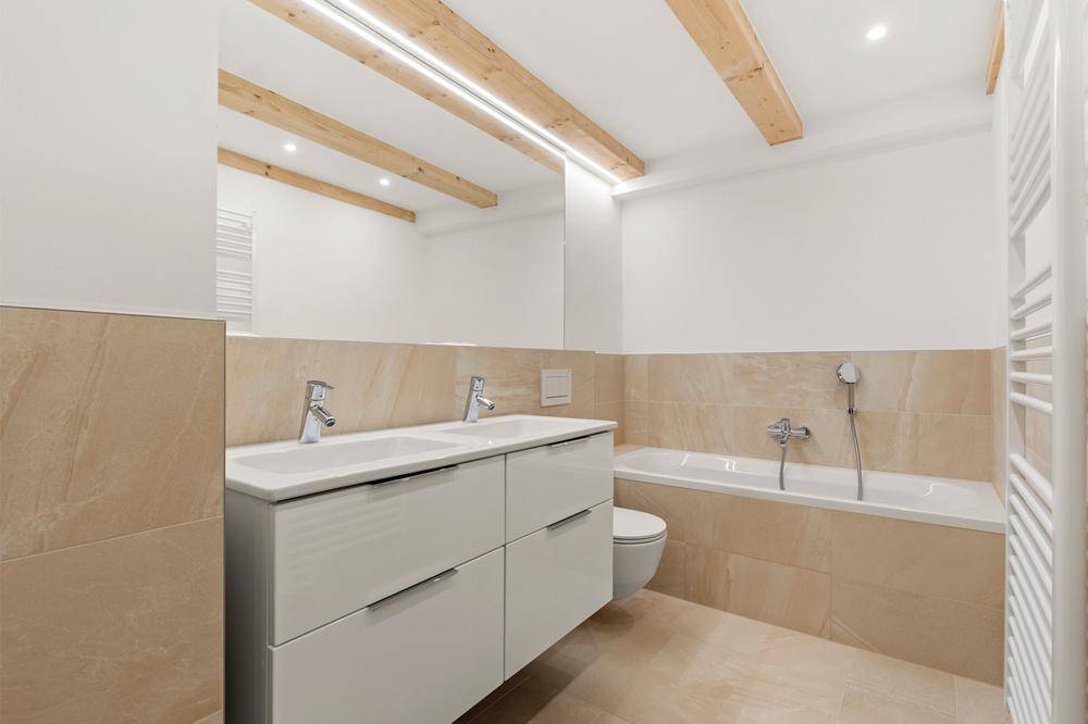 Modernes Badezimmer mit Doppelwaschbecken, Badewanne, beigen Fliesen und sichtbaren Holzbalken.