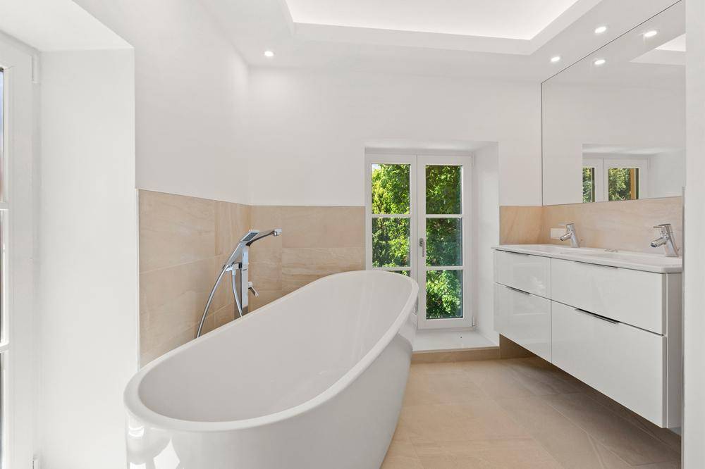 Elegantes Badezimmer mit freistehender Badewanne, Doppelwaschbecken und Fenster, in hellen Beigetönen gehalten.