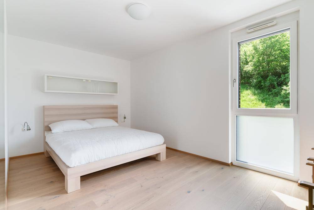 Helles Schlafzimmer mit Holzbett, weißer Bettwäsche und großem Fenster mit Blick ins Grüne.