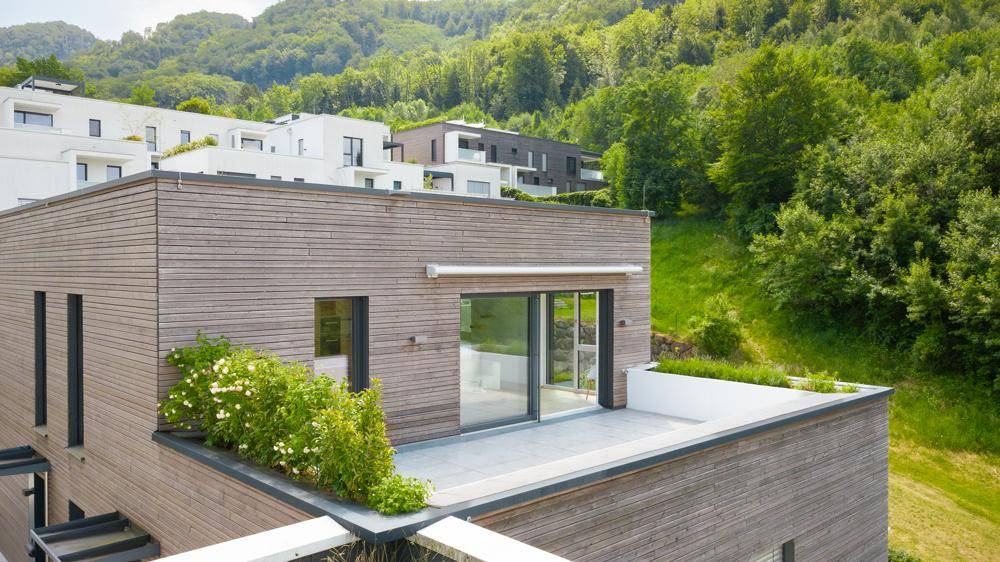 Moderne Architektur mit Holzfassade und großer Dachterrasse, umgeben von grünen Hügeln.